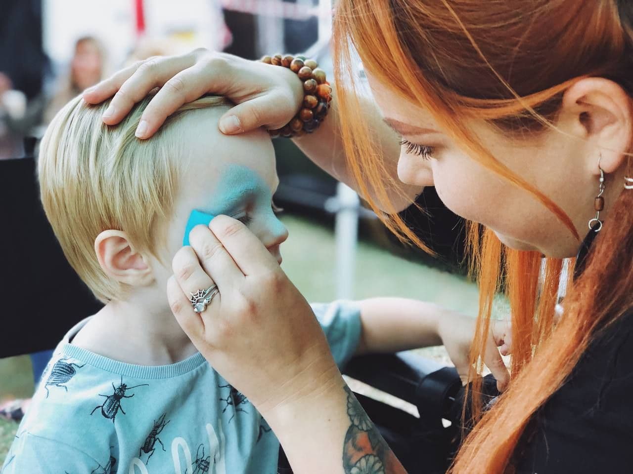 Alex applying blue face paint to a young boy at an outdoor event